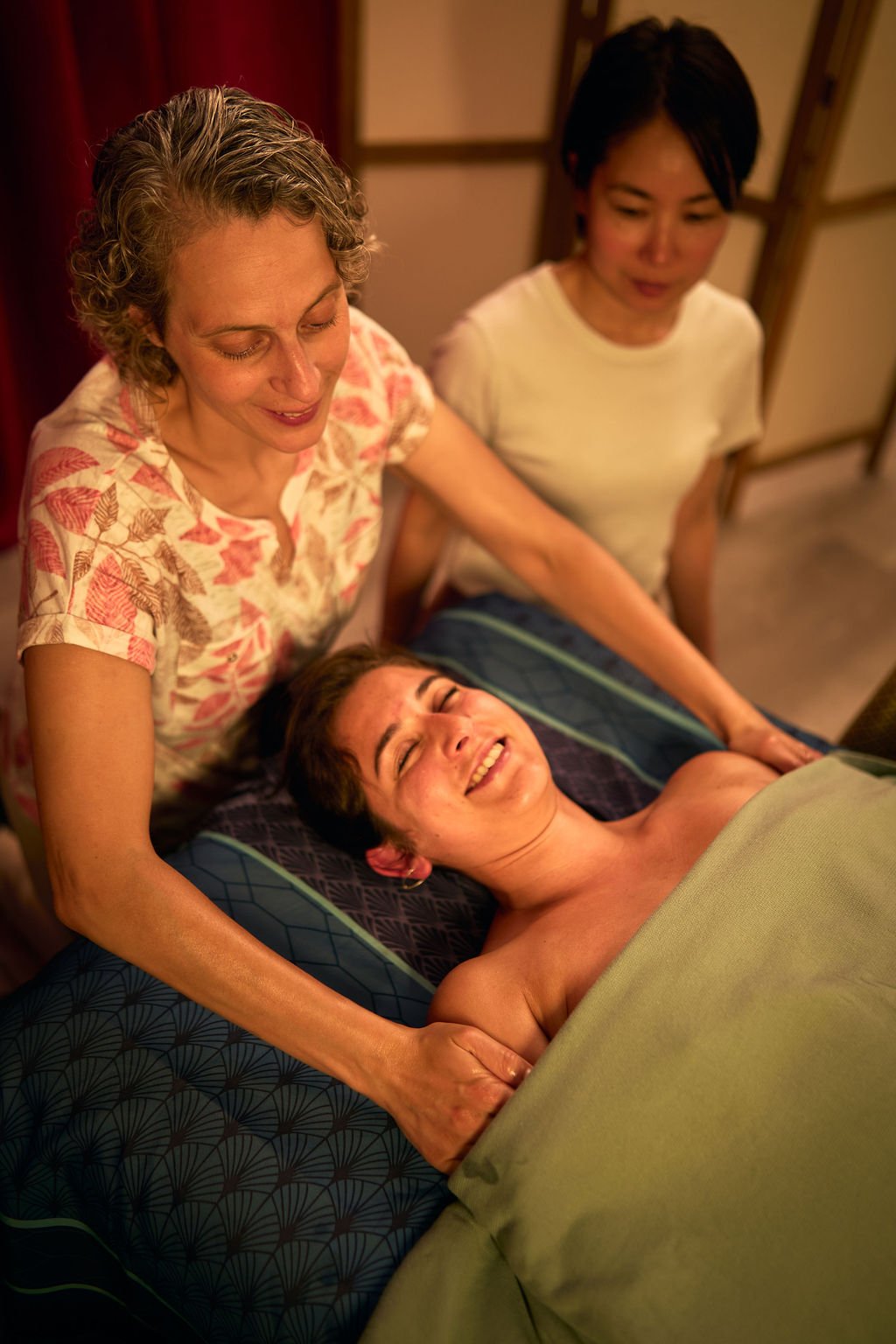 Une femme allongée dans un lit, souriant, entourée de deux autres femmes qui lui tiennent la main et lui offrent du soutien lors d'un moment de relaxation ou de massage à l'intérieur d'un environnement chaleureux.