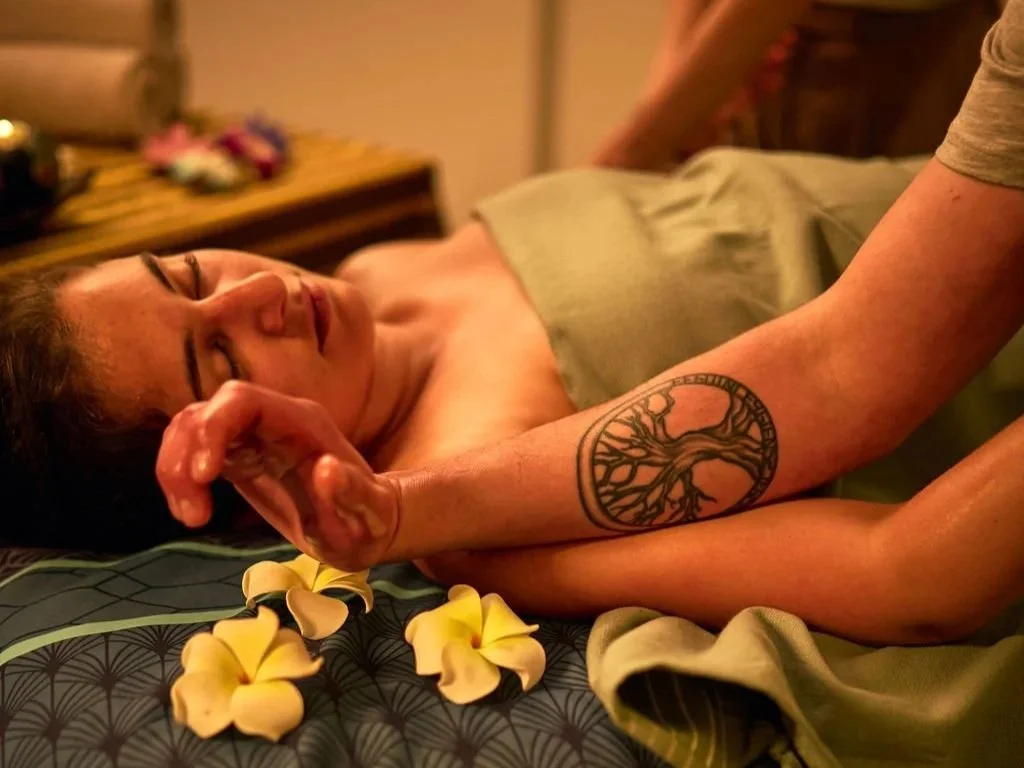 Une femme allongée dans un lit, ses yeux fermés, avec des fleurs jaunes autour d'elle et un tatouage de branche d'arbre sur son bras.