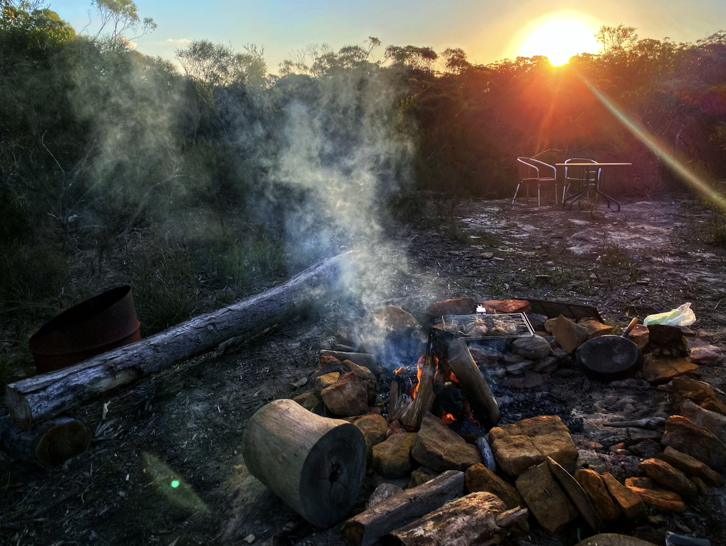 A campfire burning with smoke rising, surrounded by rocks, on a rocky outdoor area during sunset with two chairs and a table in the background.