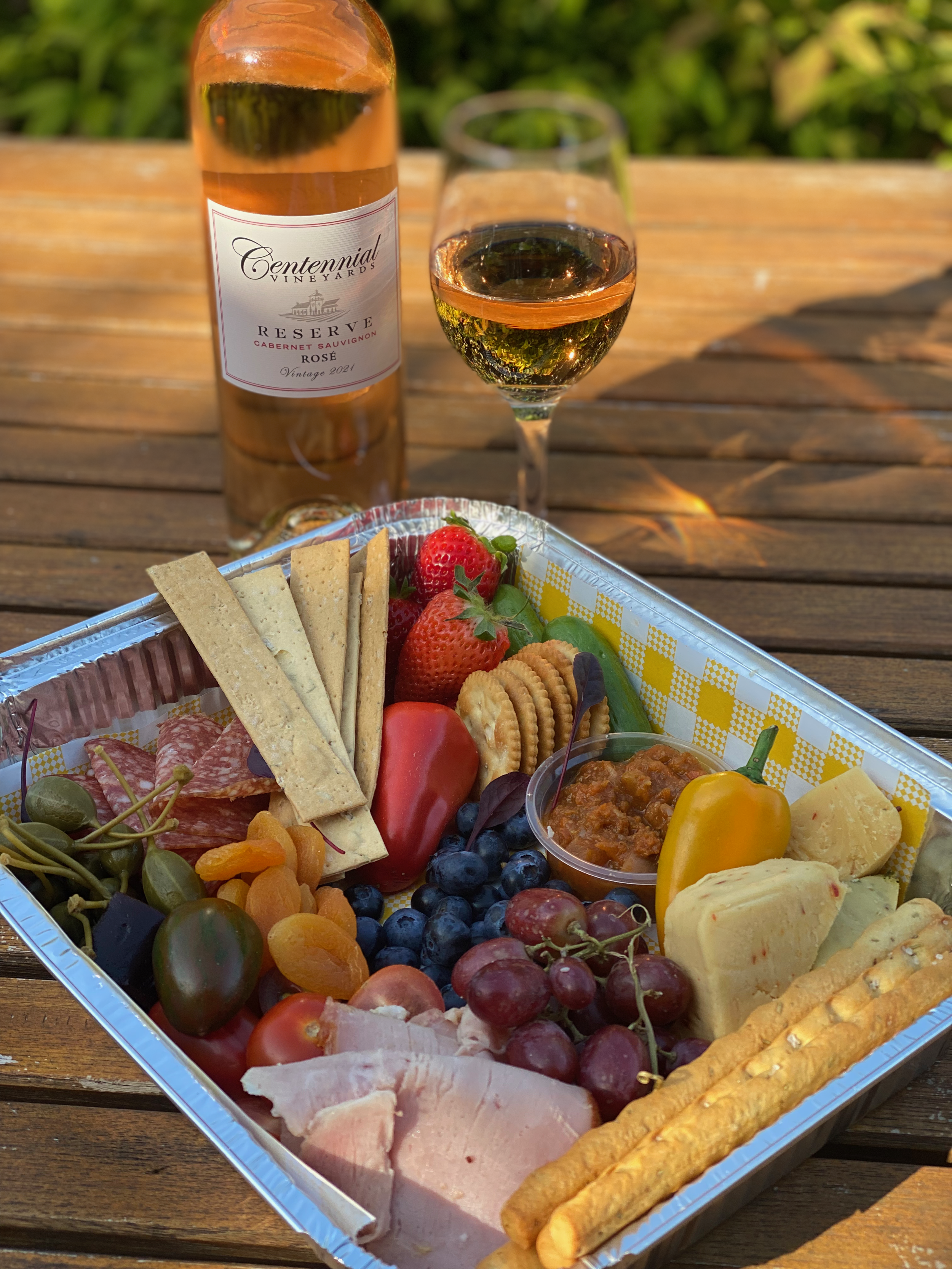 A tray of assorted fruits, cheeses, crackers, cured meats, and a bowl of spread, with a bottle of rosé wine and a glass of wine on a wooden table outdoors.