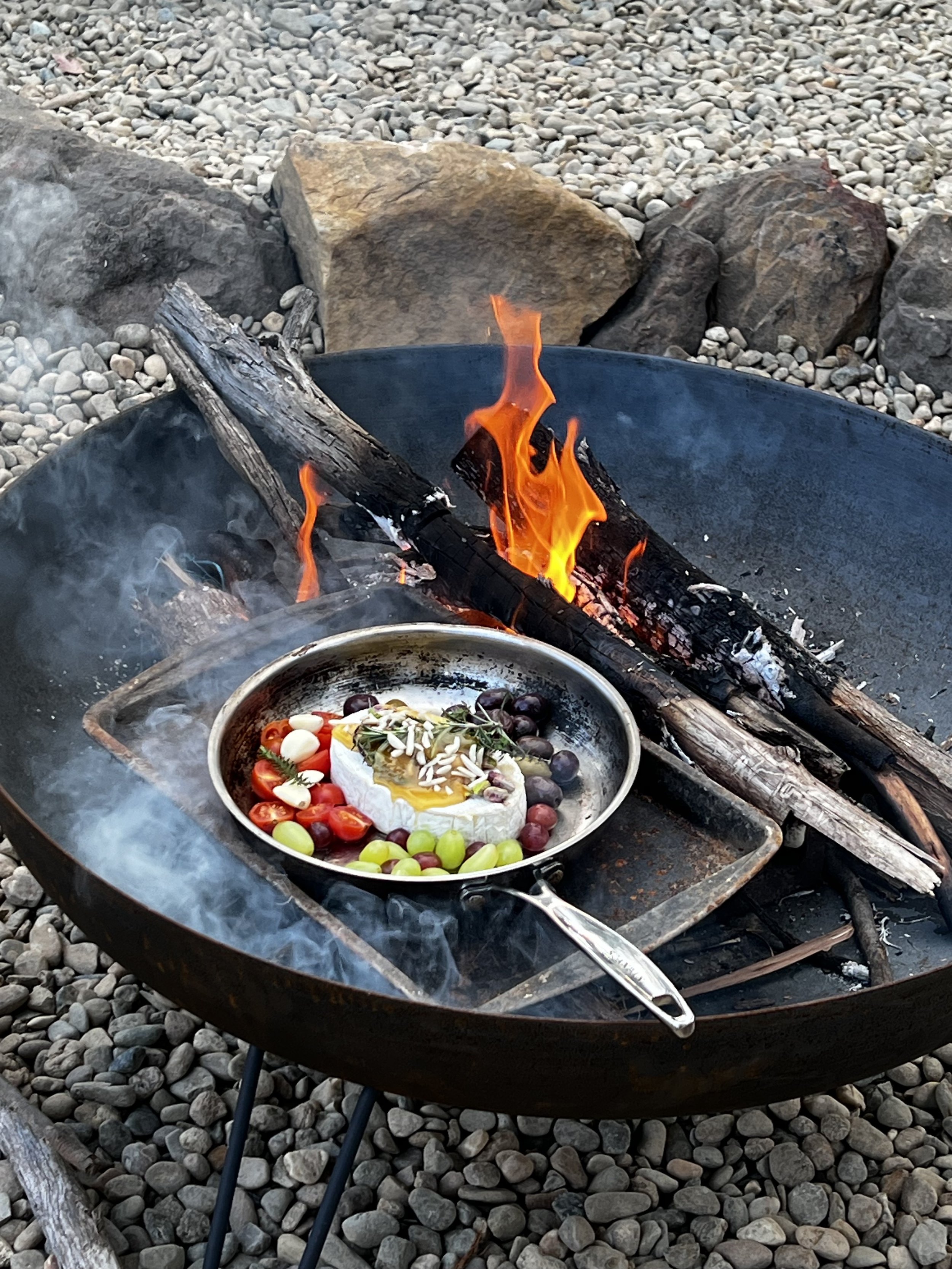 A black fire pit with a small fire, surrounded by rocks. Inside the fire pit, there is a frying pan with a dessert topped with nuts, grapes, cherry tomatoes, and a sprig of greenery. The fire pit is on a surface covered with small gray rocks.