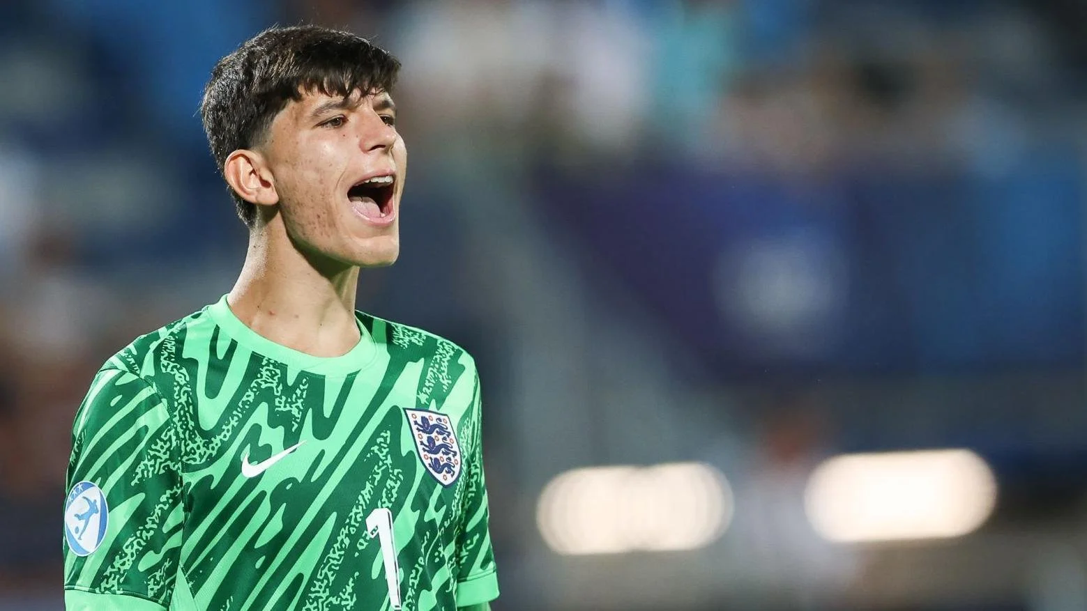 A young male soccer player wearing a green England national team jersey, shouting on a football field.