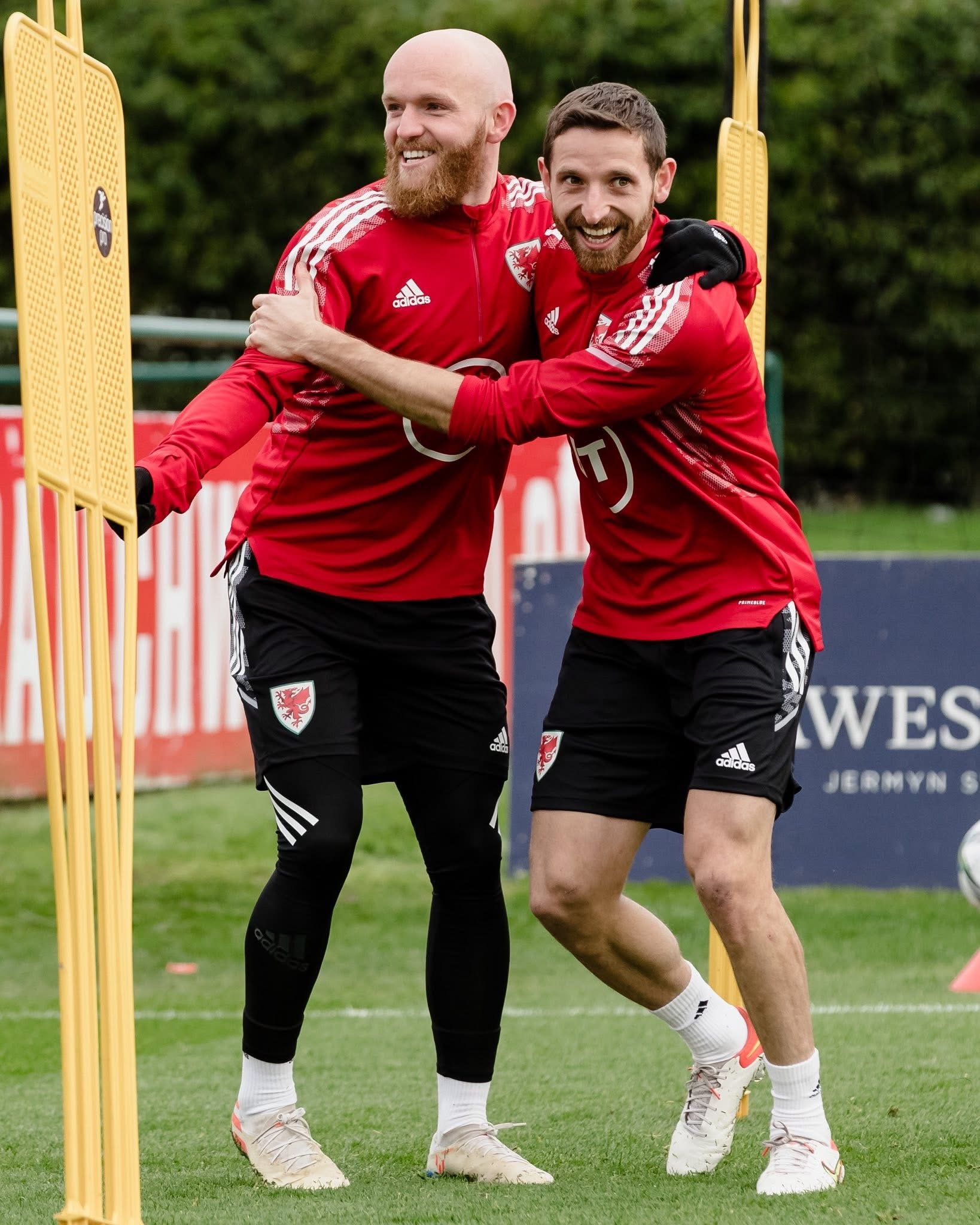 Two male soccer players wearing red jerseys with national team crests and Adidas logos, joyfully celebrating on a green field during practice, surrounded by training equipment and trees.