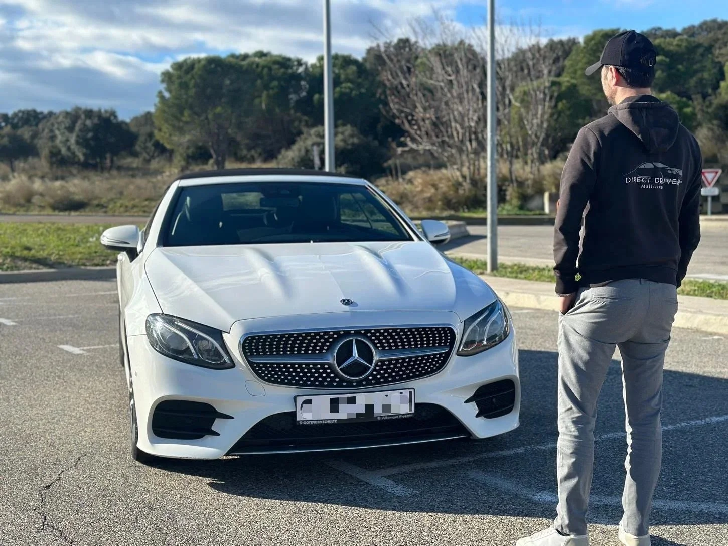A white Mercedes-Benz parked in an outdoor lot with a man wearing a black hoodie and gray pants standing nearby, facing the car. The hoodie has text and a car logo on the back. The background shows trees, a blue sky with some clouds, and a street sign.