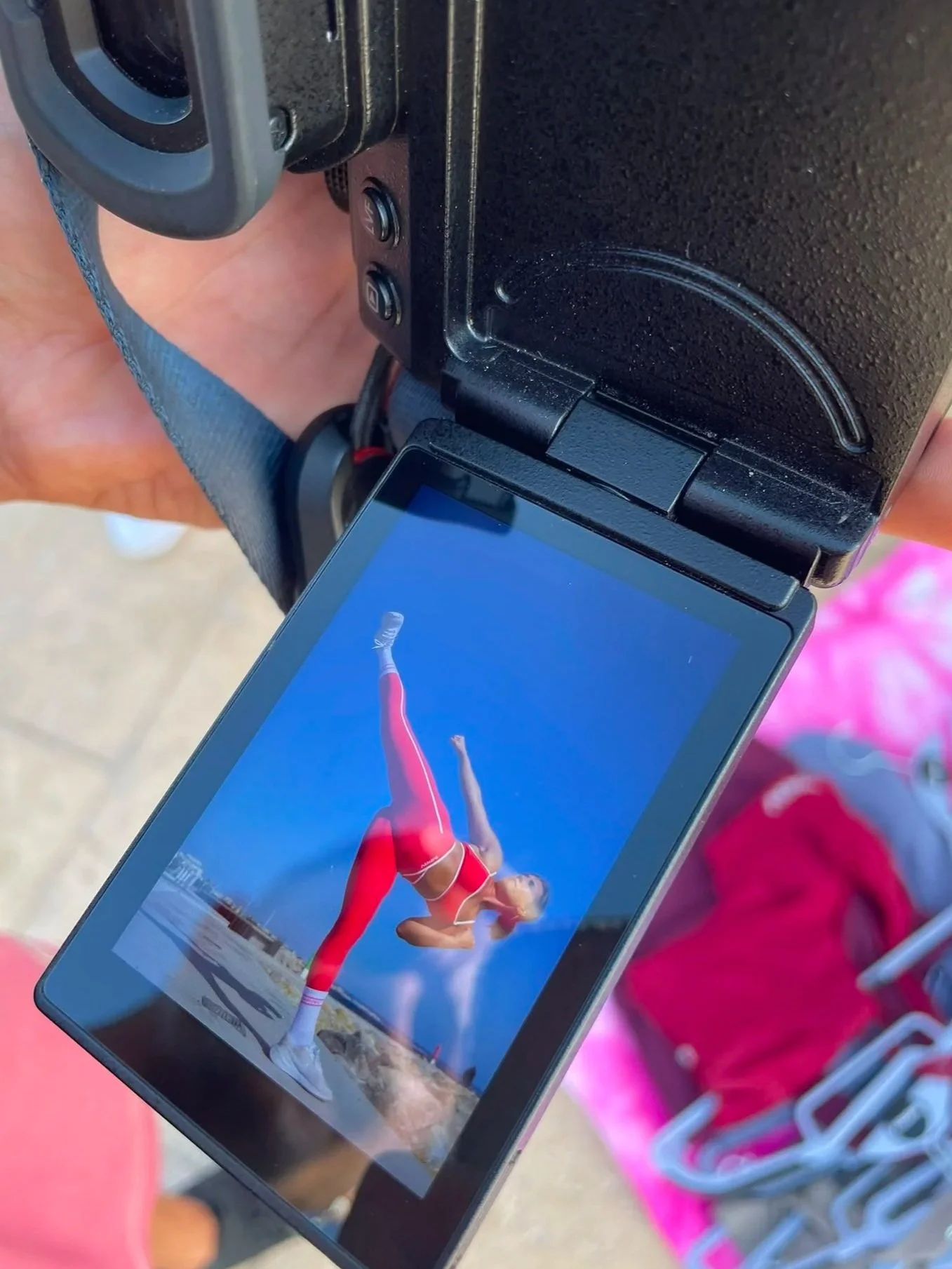 A smartphone showing a woman in a red workout outfit performing a fitness pose outdoors, held by a person with a hand visible in the background.