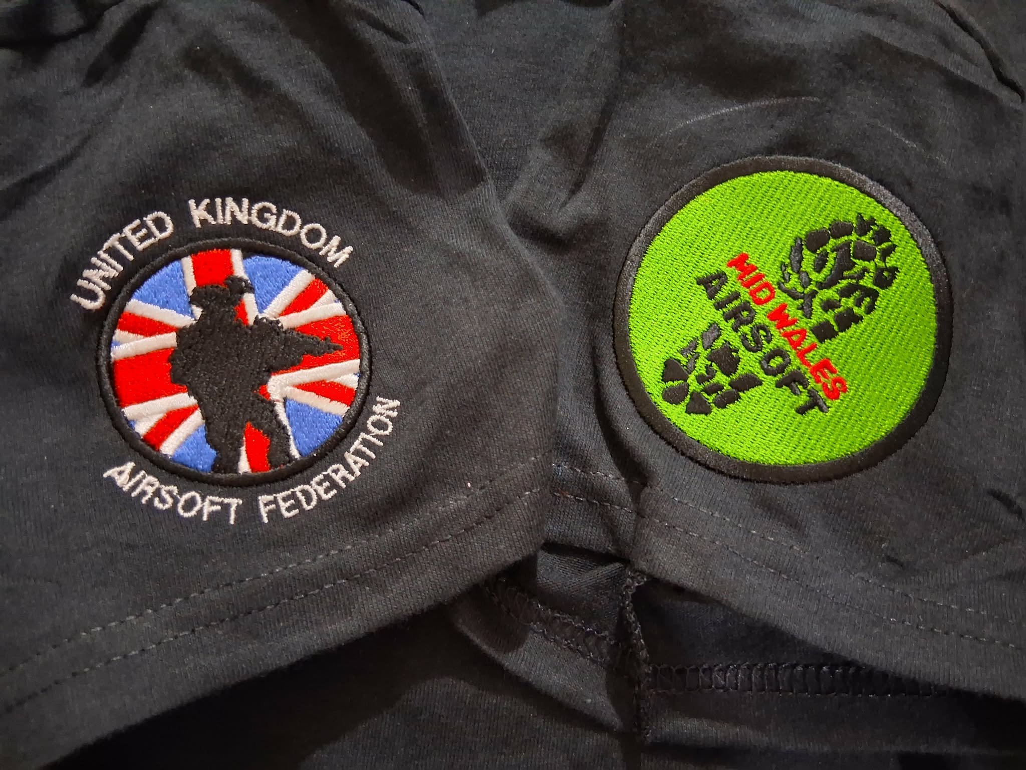 Close-up of two embroidered patches on a black fabric. One patch shows a silhouette of a dog with a Union Jack background and the words "United Kingdom" and "Airsoft Federation." The other patch is green with black camouflage pattern and red text reading "HID WARS."