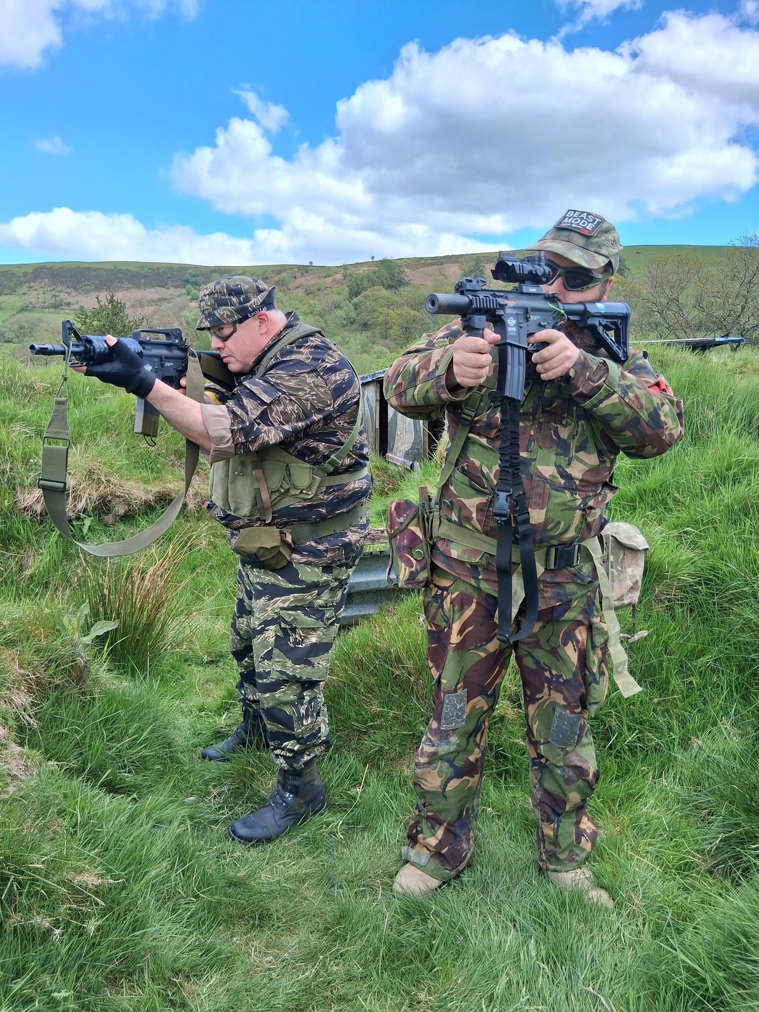 Two men in camouflage clothing aiming rifles outdoors in a grassy area with a hilly landscape and blue sky with clouds.