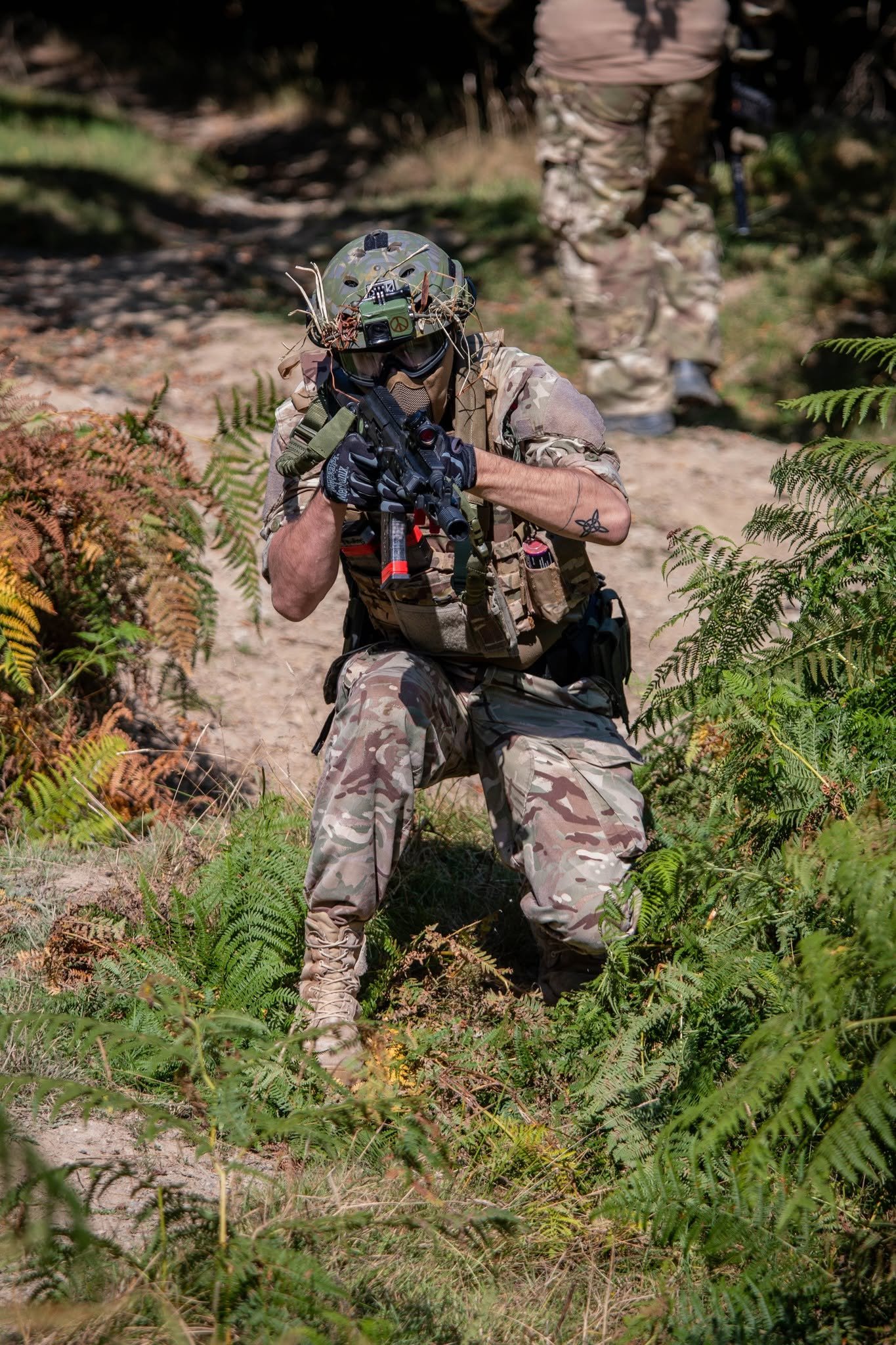 Soldier in camouflage uniform kneeling in a wooded area, wearing a helmet and tactical gear, aiming an assault rifle.