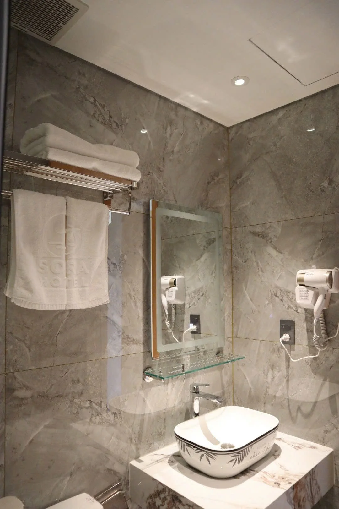 A bathroom with gray marble walls, a wall-mounted mirror, towels on a metal rack, a hairdryer, and a decorative sink on a marble countertop.
