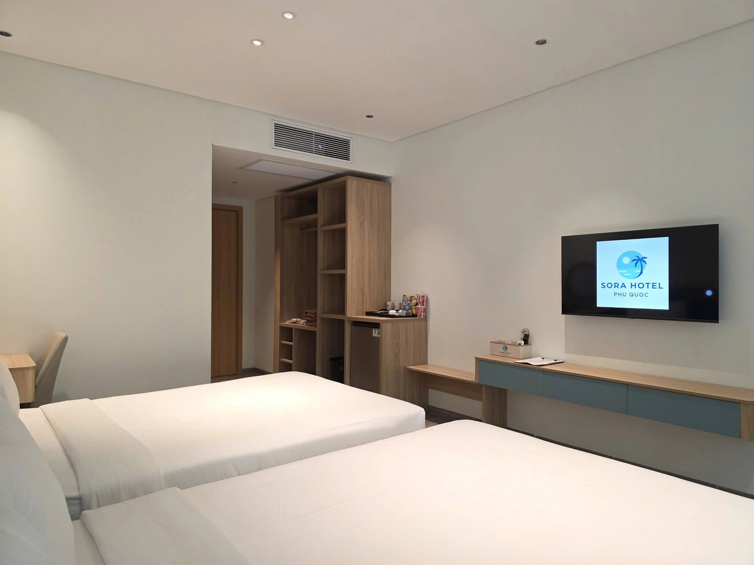Hotel room with two white beds, wooden shelving unit, television displaying Sora Hotel logo, and minimalist decor.