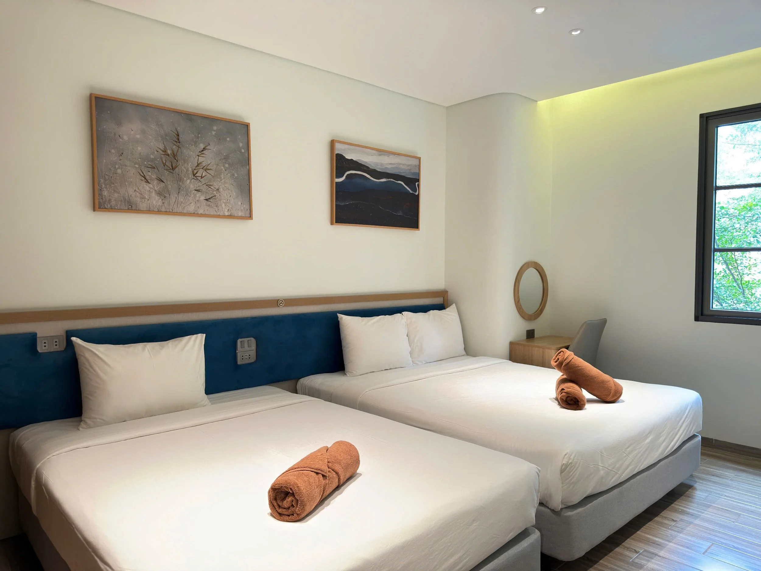 Clean hotel room with two neatly made beds, each with white bedding, pillows, and rolled brown Towels. The wall behind the beds has framed artwork, and there is a window with a view of green trees.
