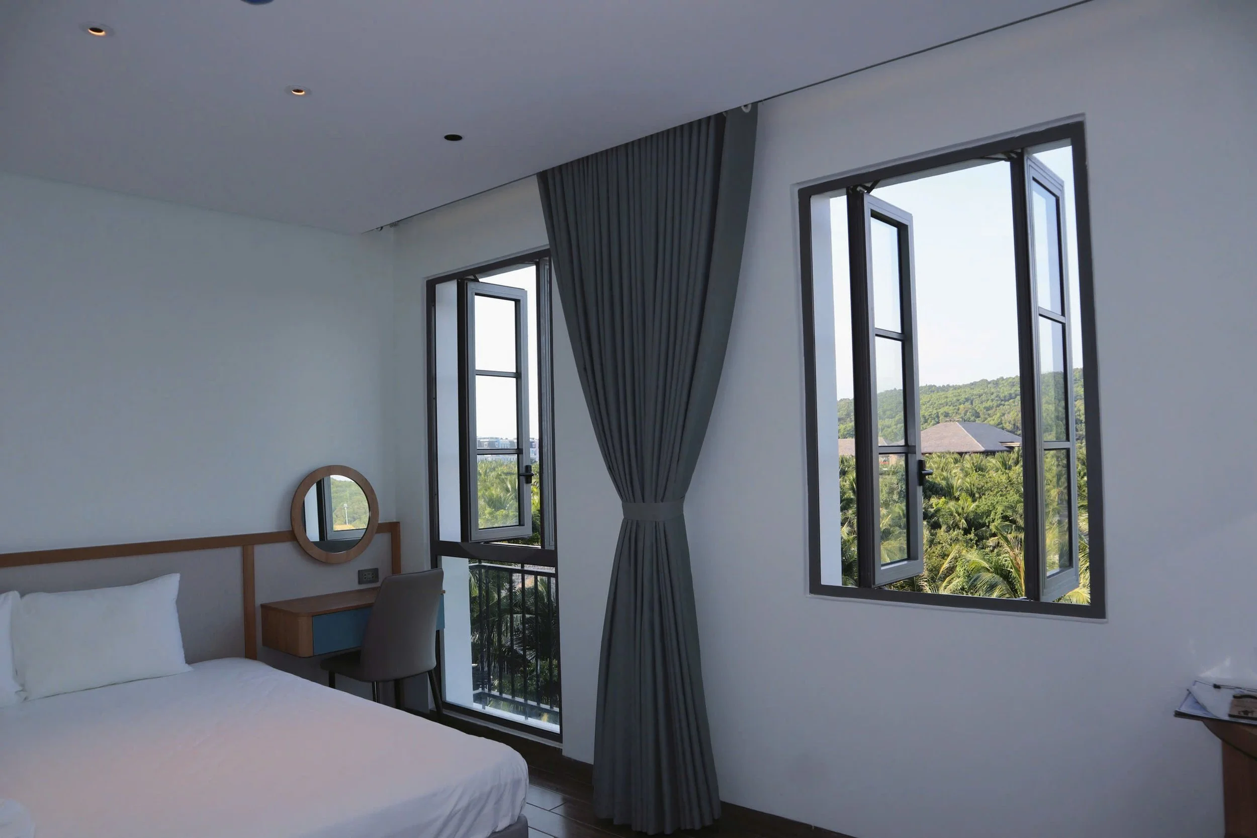 A modern hotel room with a large window showing lush greenery outside. The room has white walls, a bed with white linens, a wooden headboard, a nightstand, a vanity mirror, and a gray curtain partially open to the outside view.
