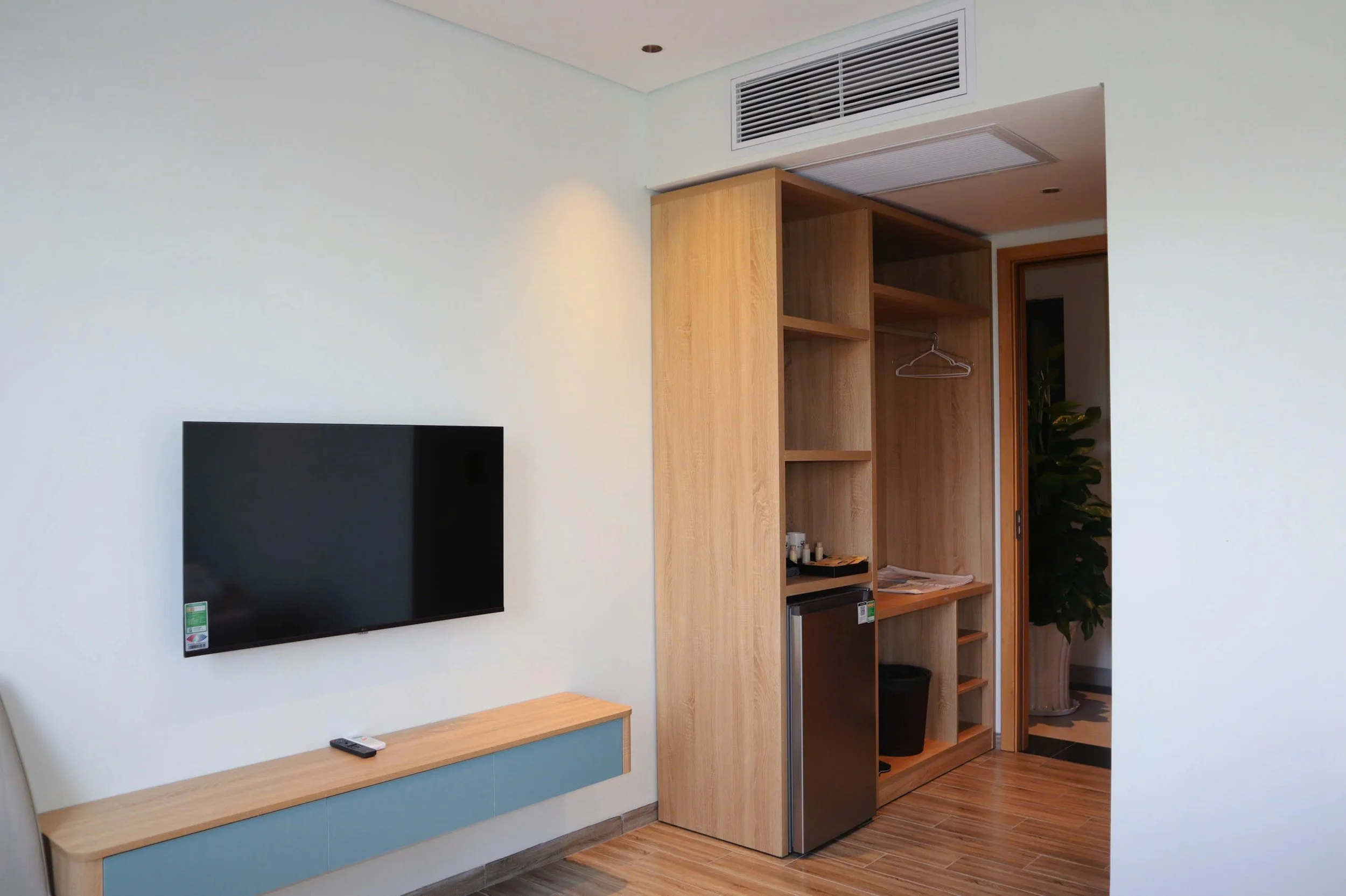 Interior of a modern living space featuring a mounted flat-screen TV on a white wall, a small wooden table below the TV with a remote control, a wooden open shelving unit with compartments, a built-in mini refrigerator, and an open doorway leading to another room with a large plant.