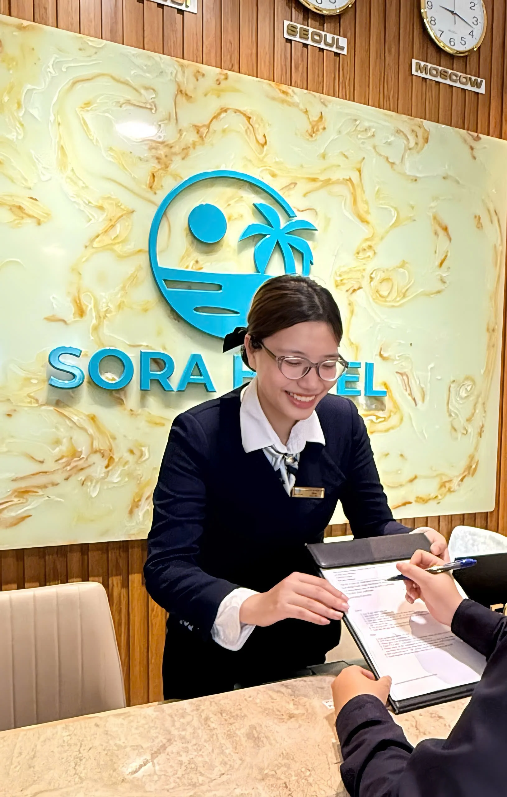 A woman smiling and wearing glasses, sitting at a table, holding a menu or document, in front of a sign with the logo and name 'Sora R'cel' and a background wall with clocks showing different times and signs indicating 'Seoul' and 'Moscow'.