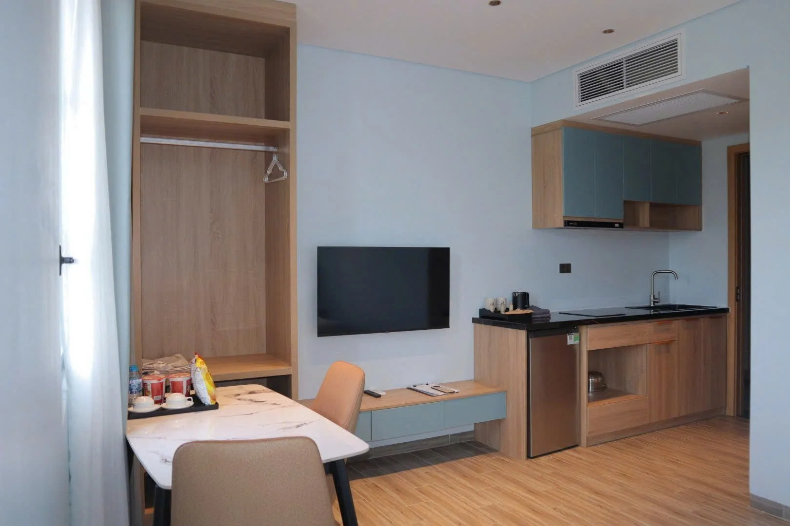 A modern hotel room kitchenette with a small black countertop, a sink, wooden cabinets, a mounted flat-screen TV, a dining table with beige chairs, and a partial view of a wardrobe with an open space and hanging rod.