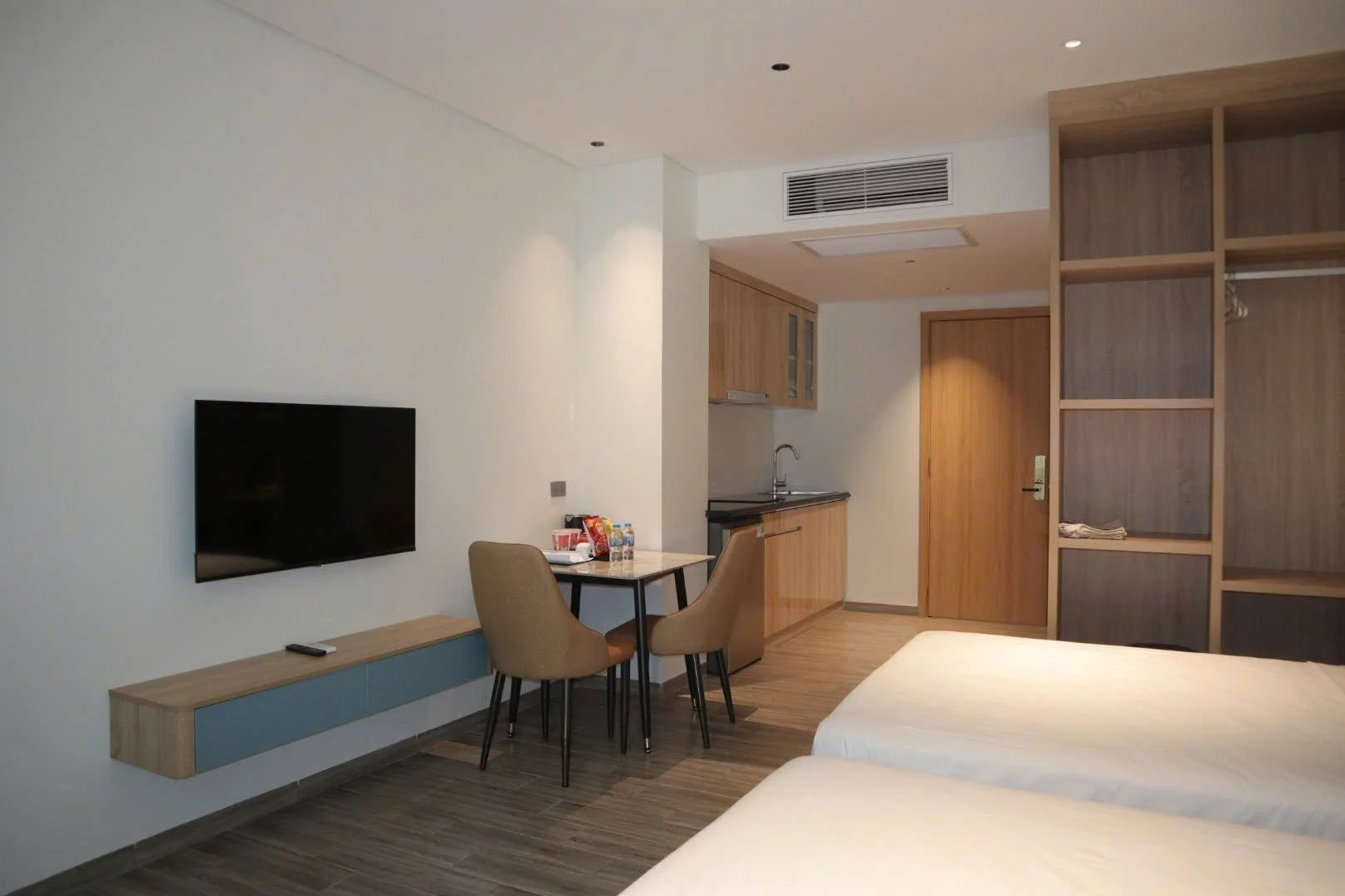 Interior view of a modern hotel room with a small dining area, kitchenette, flat-screen TV, and partial view of beds.