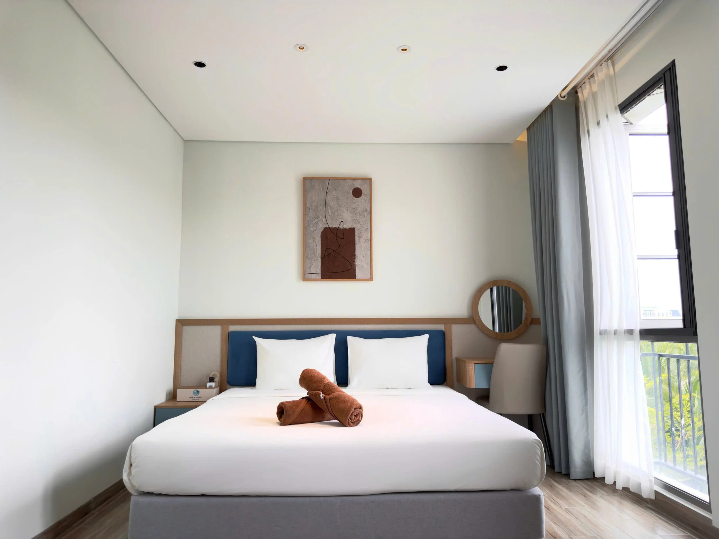 A modern hotel room with a large bed covered in white bedding and a rolled brown towel on top. The bed has a wooden headboard with a blue cushioned panel. There are two white pillows on the bed. To the left of the bed, a bedside table with a reading light, a phone, and a notepad. The room has a large window with sheer white curtains and gray drapes, allowing natural light to fill the space. A round mirror hangs on the wall next to the window, and a piece of abstract artwork is mounted above the headboard. The ceiling is plain white with recessed lighting.