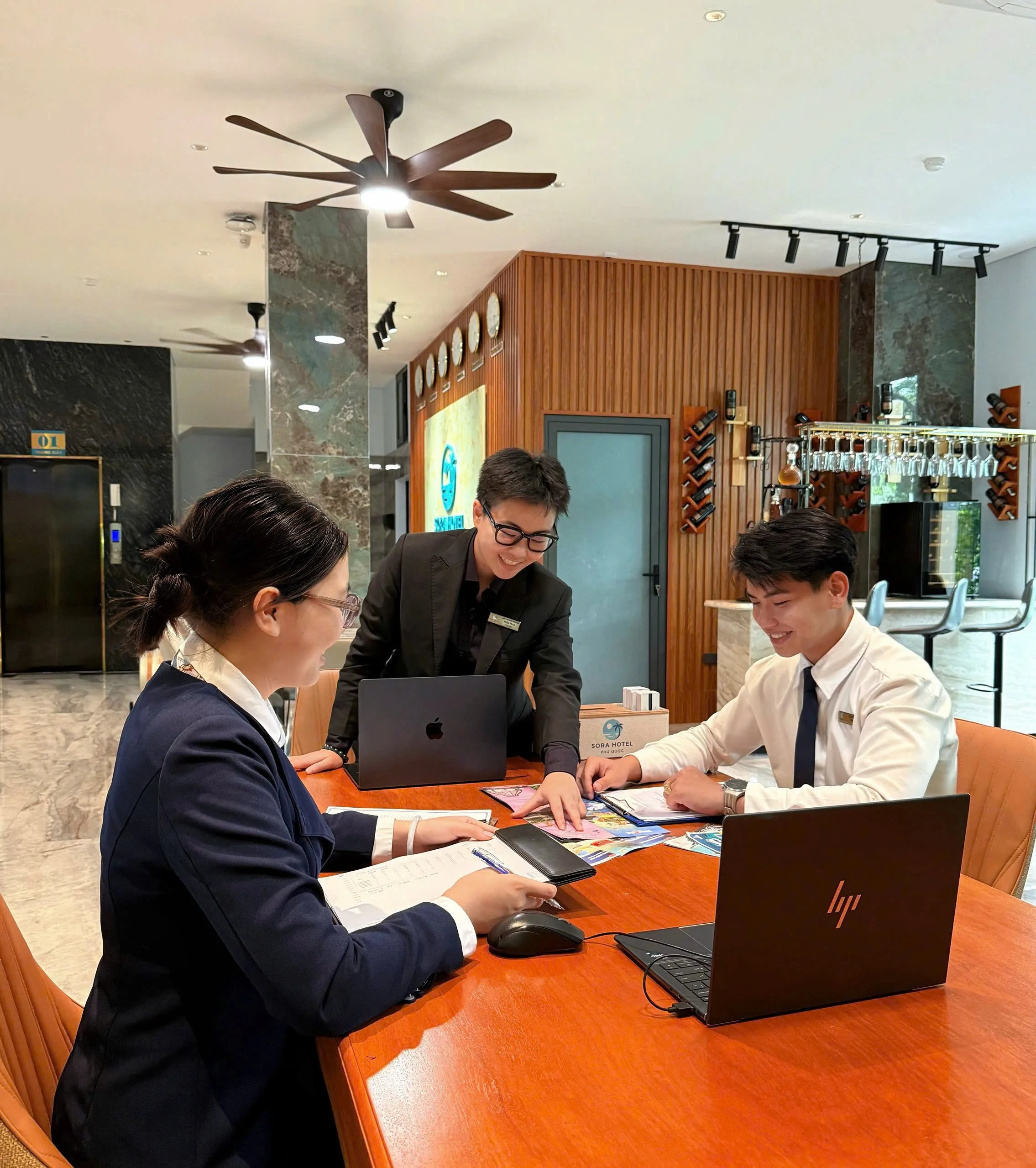 People sitting at a wooden table in a hotel lobby, engaging in discussion with documents, laptops, and brochures. A woman with glasses and a navy blazer is in the foreground, and a woman and man are smiling and interacting across the table.