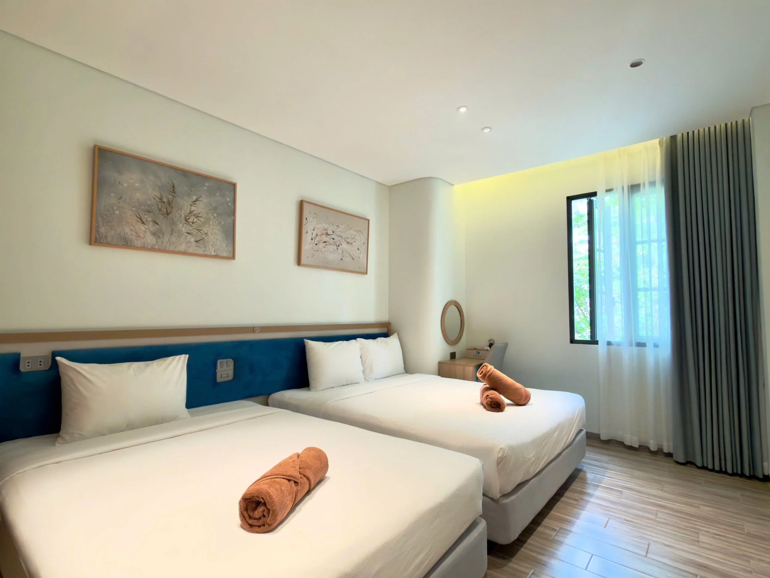 A bedroom with two single beds placed side by side, each with white bedding and pillows. Two rolled towels are on each bed. The wall behind the beds has artistic paintings. There is a window with curtains allowing natural light into the room.