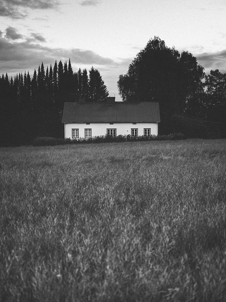 Black and white photo of a house with a sloped roof, surrounded by trees and grass, under a cloudy sky.