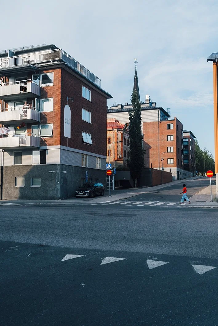 Kuvassa on kaupunkinaapuri, jossa on kerrostaloja ja kirkontorni taustalla. Ristikkäin raitiovaunupysäkki ja kävelytie, jossa on ihmisiä kävelemässä.