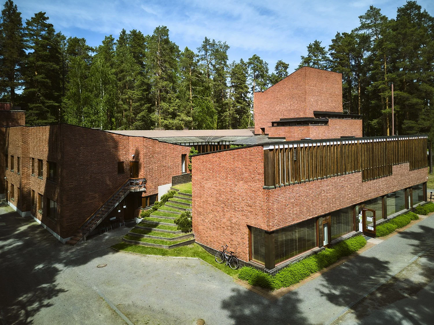 A modern brick apartment building surrounded by trees, with a small green lawn, stairs, bicycles, and large windows.