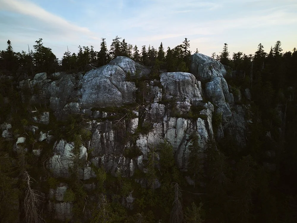 Rock formation with large boulders and pine trees, forested landscape under a partly cloudy sky.