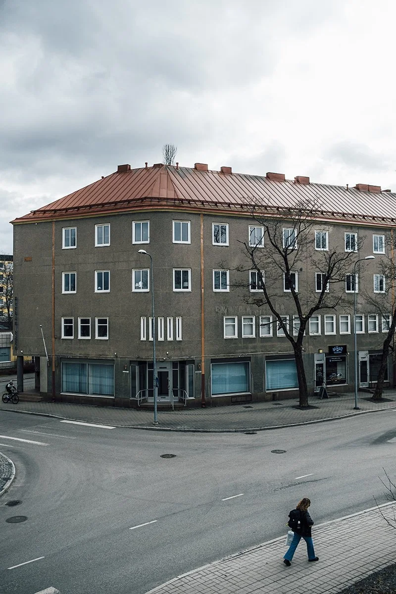 A multi-story building with a rounded corner, reddish metal roof, and multiple windows. There are leafless trees and street lamps in front, with a person walking on the sidewalk and a cyclist on the street. Overcast sky above.