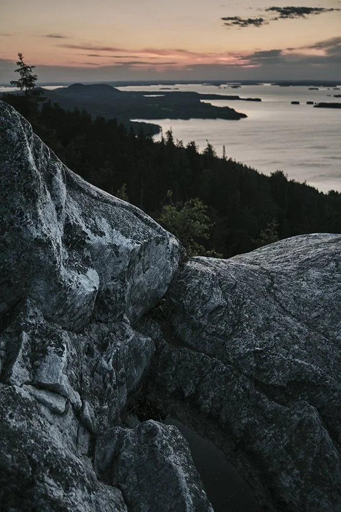 A scenic view from a rocky outcrop overlooking a body of water with islands and a forested coastline during sunset.
