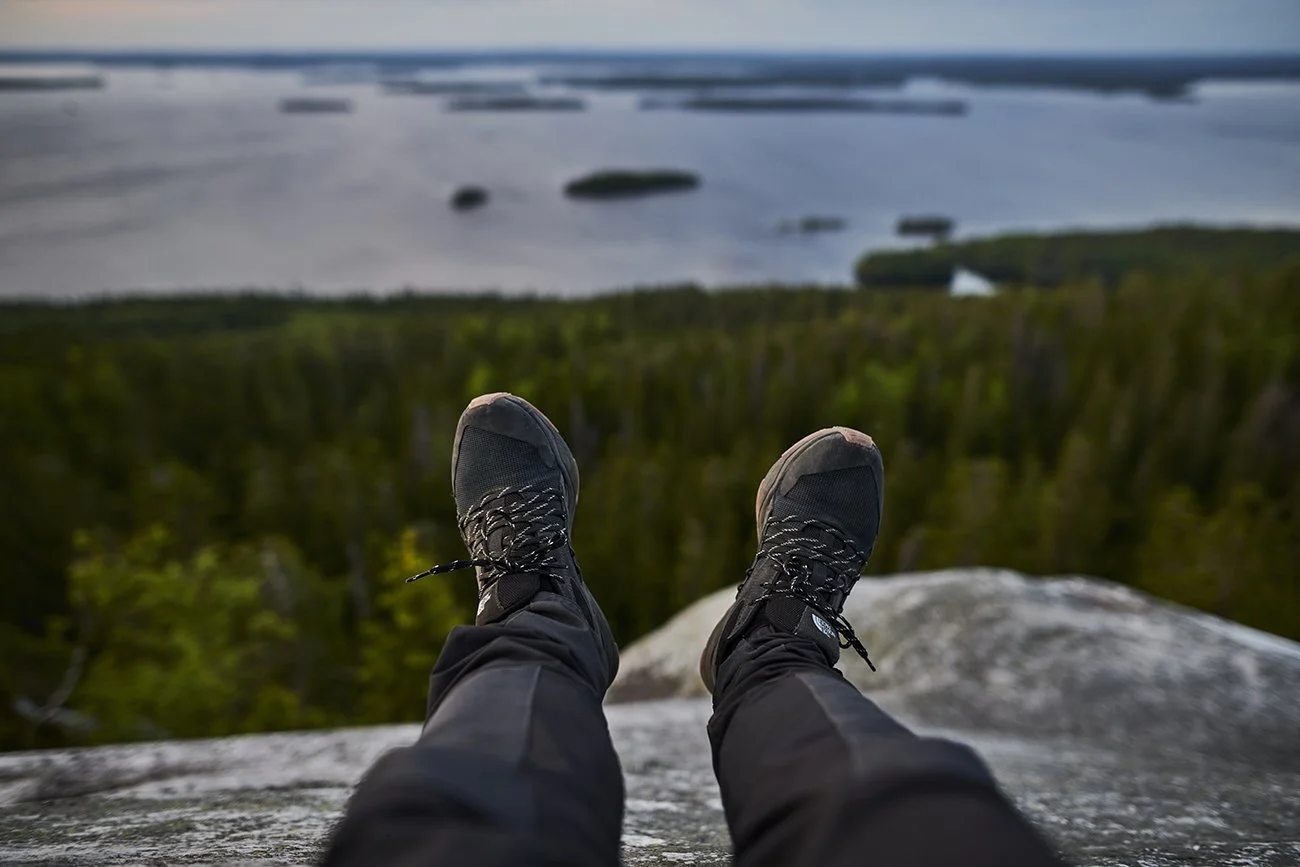 Person in black hiking boots and pants sitting on a rock ledge overlooking a landscape with trees, water, and islands in the distance.