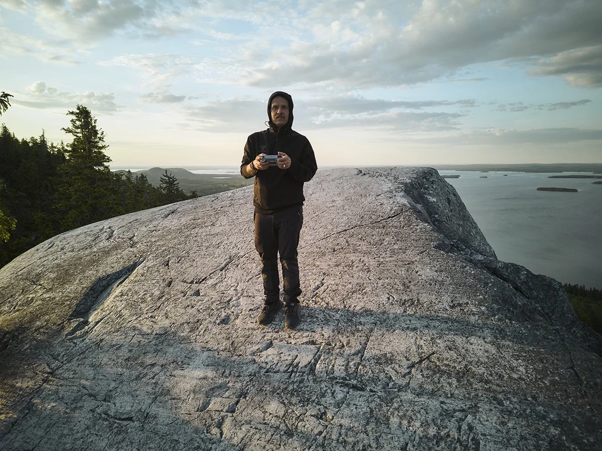 A man standing on a large rock formation on a mountain, holding a camera, with forest and water bodies in the background during sunset.
