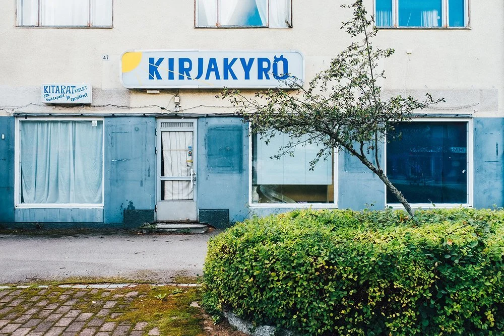 A storefront with blue painted exterior, a door, large glass windows, a sign reading 'KIRJAKYRÖ,' a small sign in Finnish, a small tree and bush in the front.