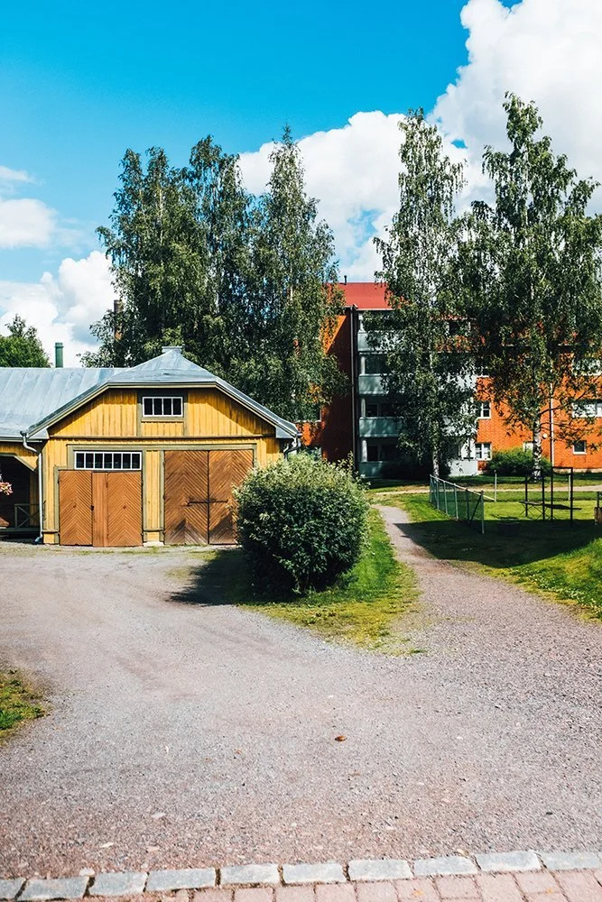 A gravel driveway leading to a wooden barn with large double doors, surrounded by green grass, trees, and a modern red-brick apartment building in the background under a partly cloudy blue sky.