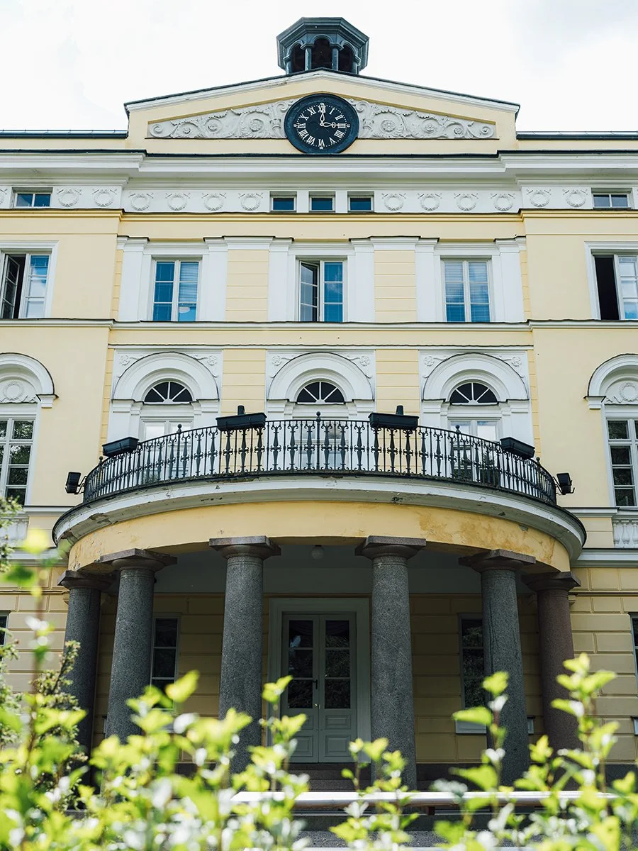 A historic yellow building with a clock at the top, a balcony with a black wrought iron railing, tall grey stone columns at the entrance, and several windows.