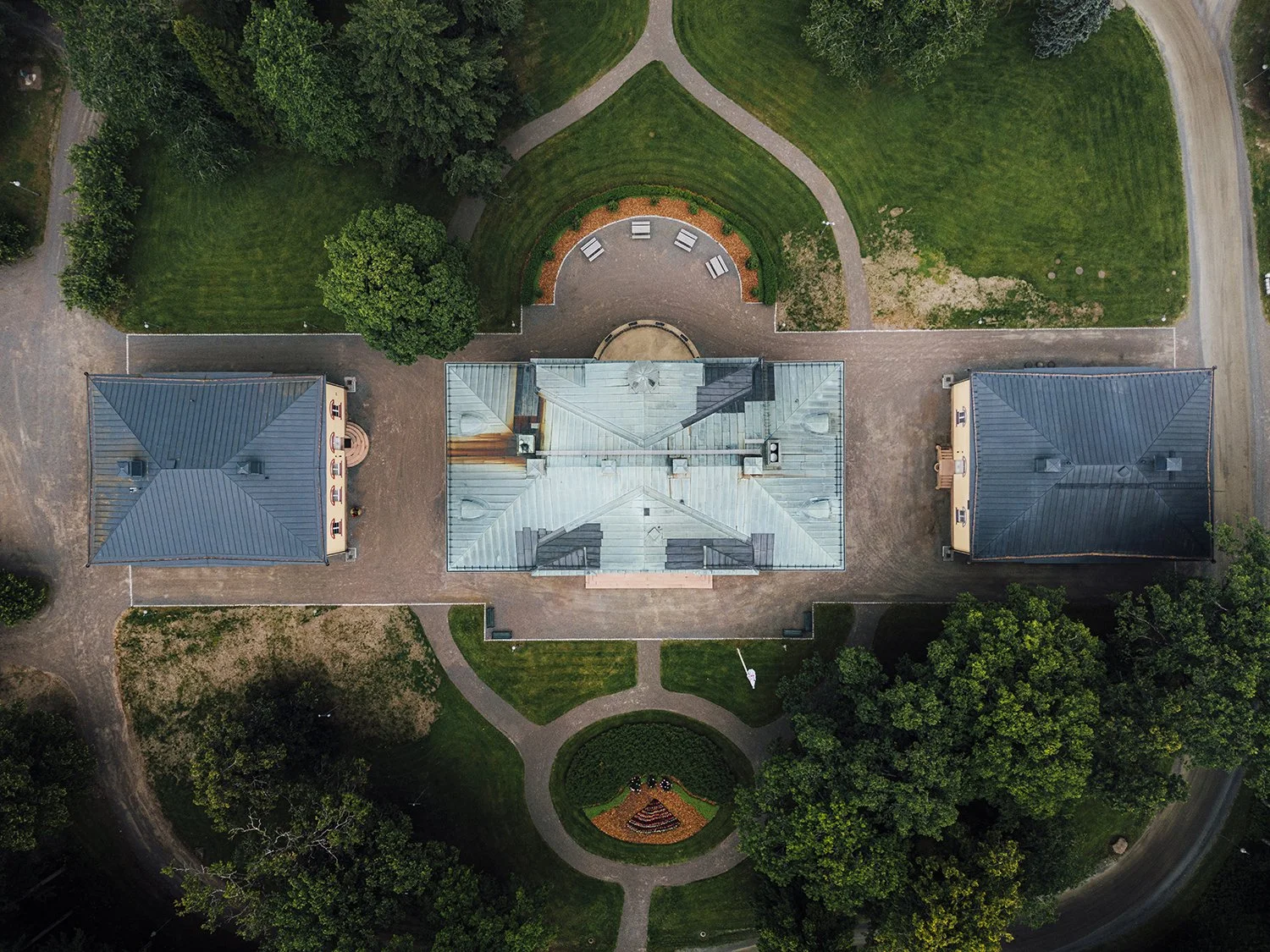 An aerial view of a large historic building with multiple wings, surrounded by green lawns, trees, and walking paths. There are two smaller buildings with blue roofs on either side of the main building. A circular garden with a flower bed is in front of the building, and benches are visible in the background.