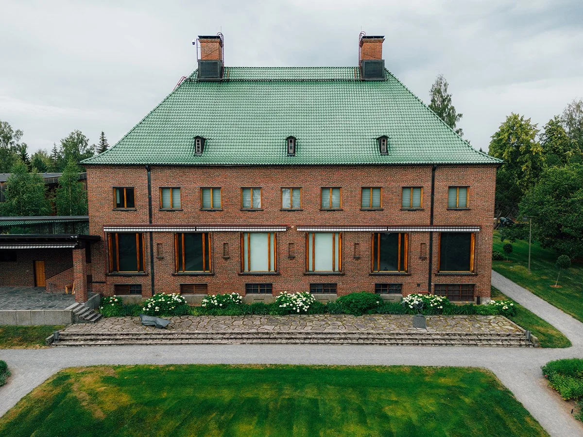 An old brick building with a green tiled roof and multiple windows, surrounded by a garden with flowers and greenery.