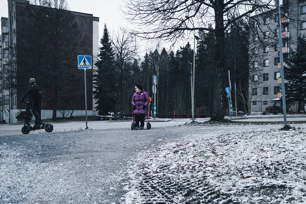 Two children with scooters in a snowy urban area, surrounded by trees, tall buildings, and street signs.
