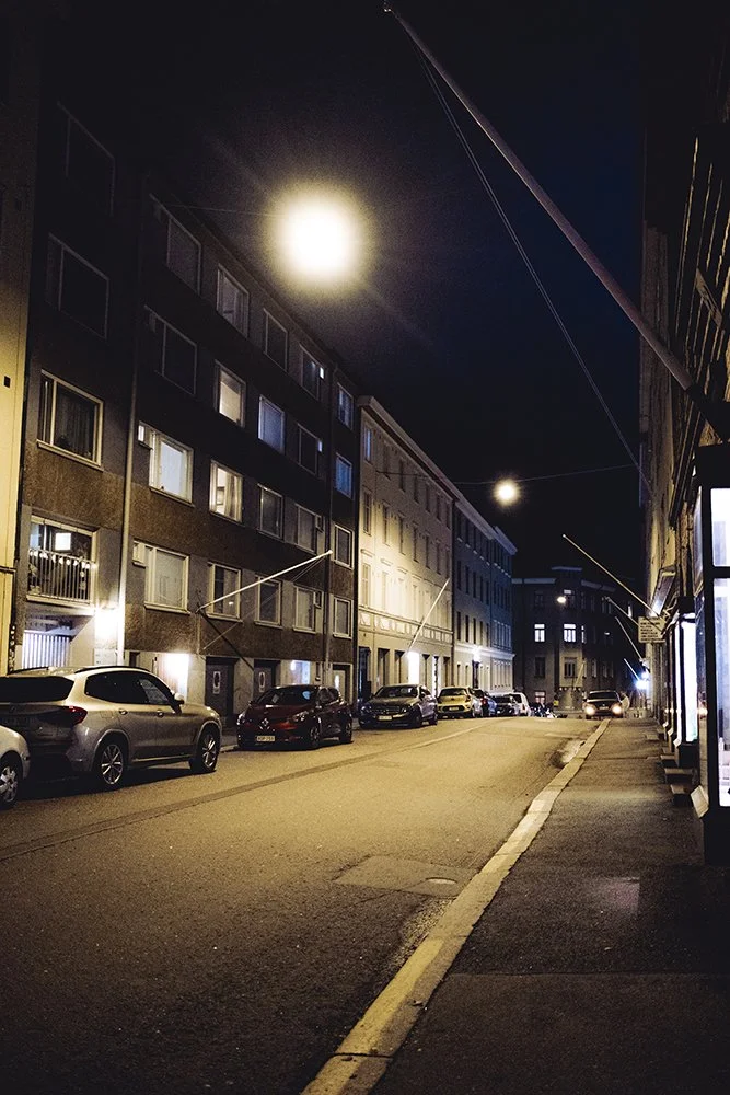 Nighttime city street scene with parked cars, multi-story buildings, and bright streetlights under a dark sky.