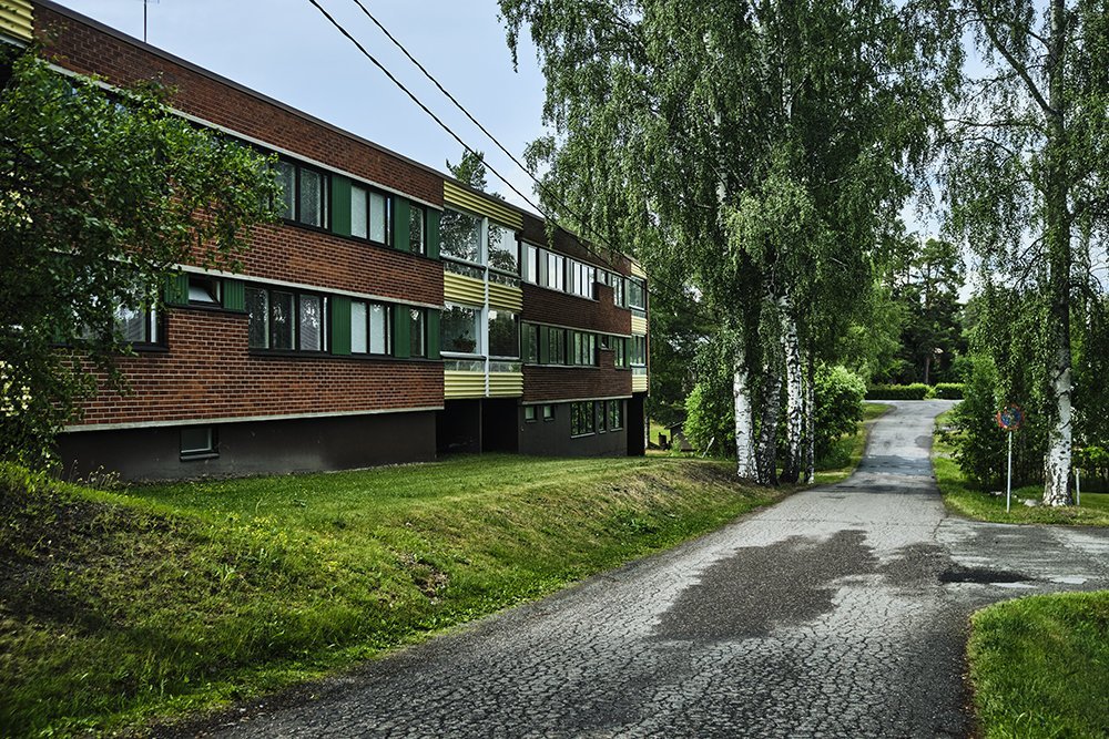 A multi-story brick residential building next to a paved, winding road with trees and greenery surrounding the area.