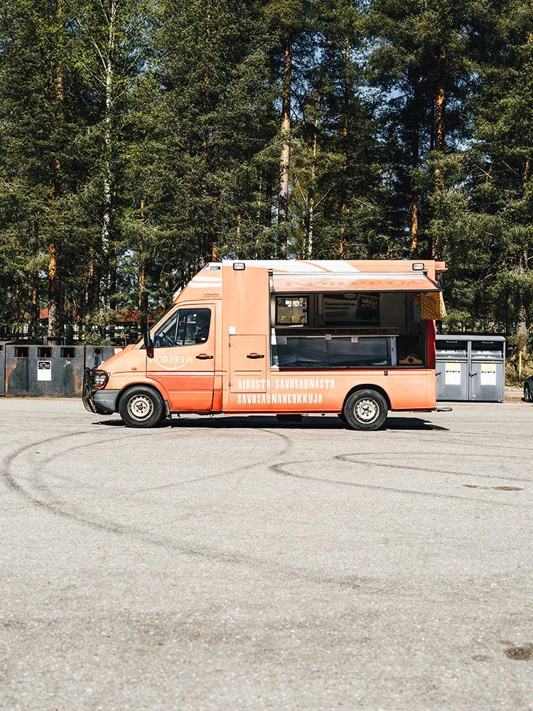 Saapastuoreauto, jossa on myyntikojut, pysäköity iso avara alueen keskelle, taustalla puita ja passiivinen taivas.