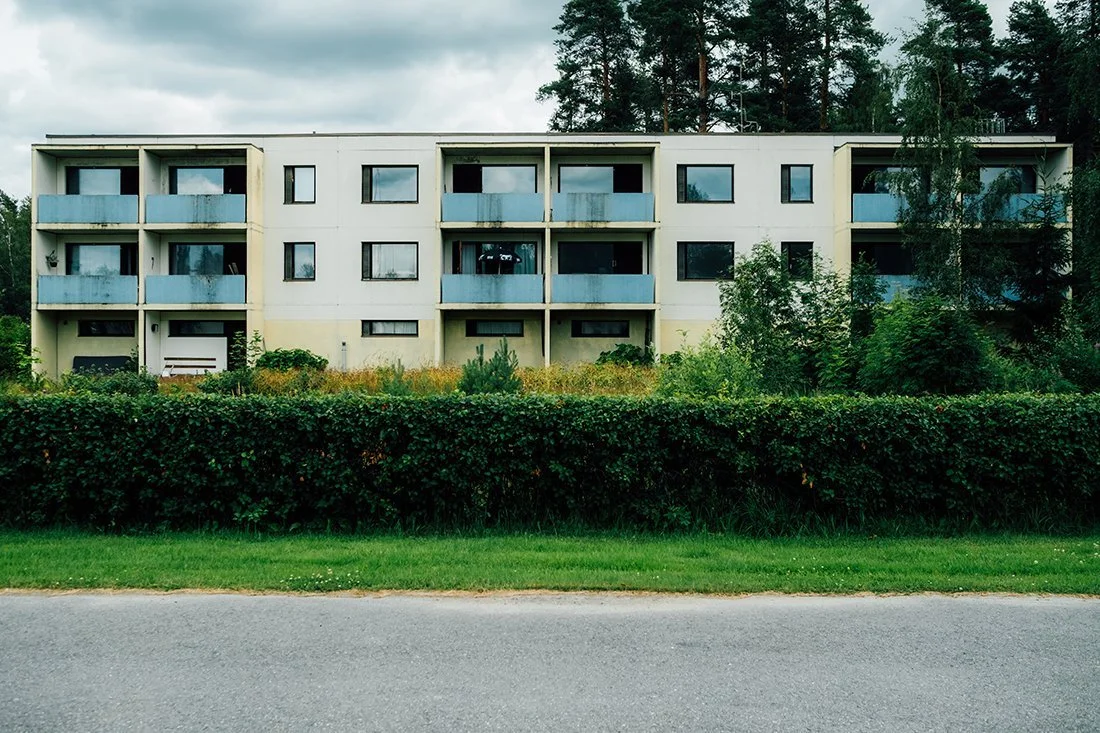 A three-story apartment building with balconies, surrounded by greenery and trees, under a cloudy sky.