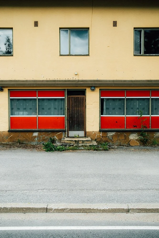 A two-story building with beige exterior walls, two upper windows, and two large ground-floor windows with red and gray panels. There is a wooden door between the ground-floor windows, and small plants grow on the sidewalk near the foundation.