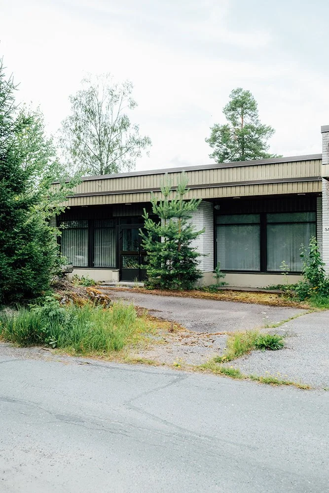 A single-story commercial building with large glass windows, front door, surrounded by trees and greenery, with a paved parking area.