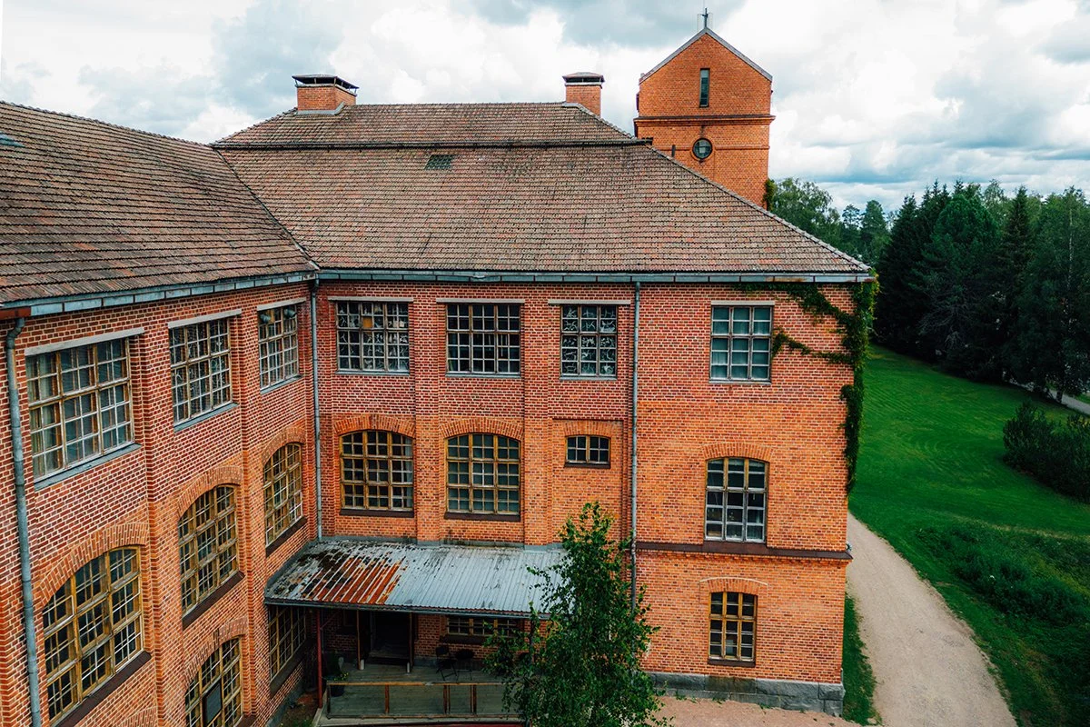 A historic red brick building with a sloped tiled roof, multiple large windows, and a small covered patio with a corrugated metal roof, surrounded by green grass and trees under a cloudy sky.