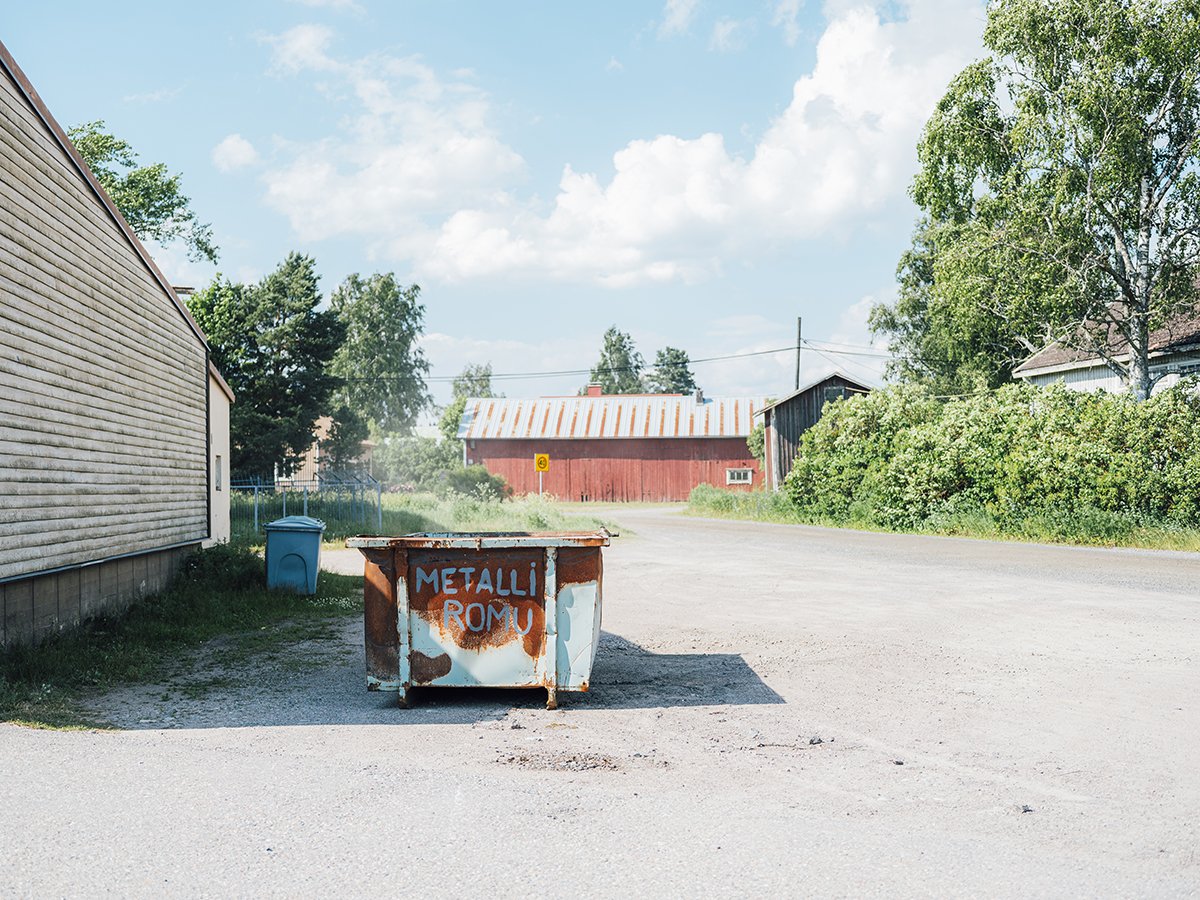 Ränsistynyt metalliastia kyltti, jossa lukee "METALLI ROMU", sijaitsee hiekkatiellä, taustalla puita ja maalaistaloja, sininen roskapönttö ja kirkas taivas.