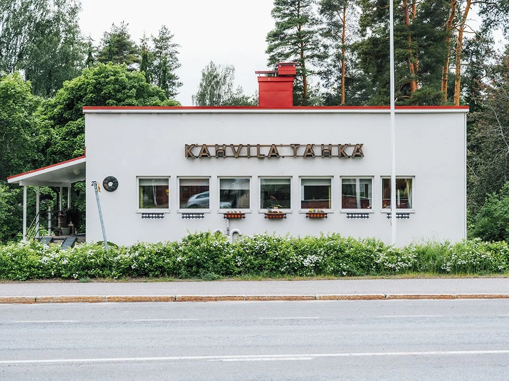 Valkoinen kahvila rakennus, jossa on teksti "KAHVILATAKKA", ympäröi vihreitä puita ja pensaita.