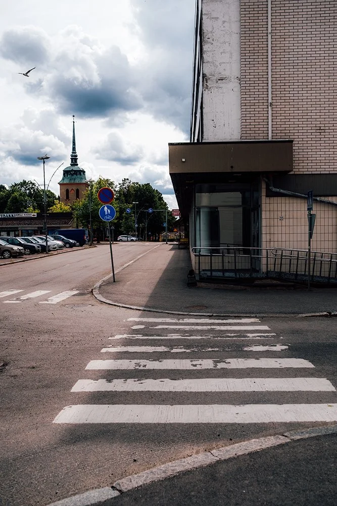 Empty crosswalk on a city street with parked cars, a no-parking sign, and a church steeple in the background under cloudy skies.