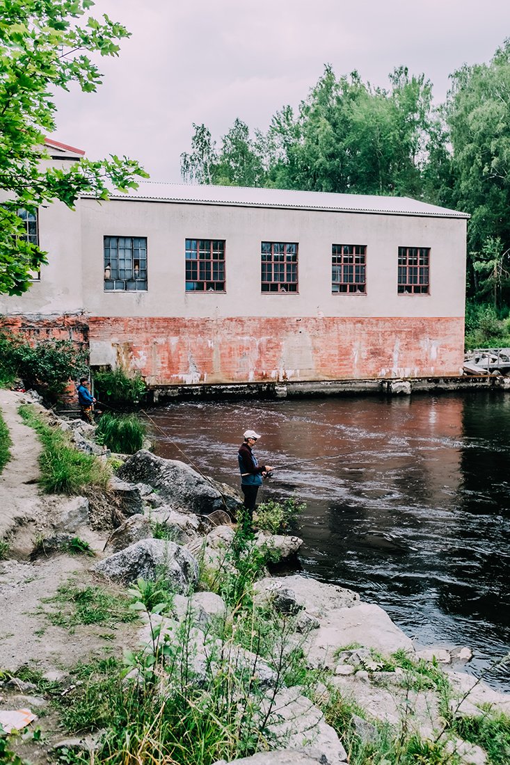 Kaksi ihmistä kalastamassa joen rannalla, taustalla teollinen rakennus ja puita