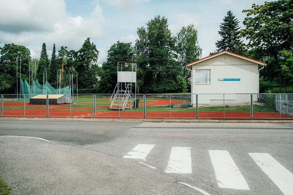 An outdoor athletics field with a running track, a white building, and a tall referees' tower, surrounded by a metal fence and trees.