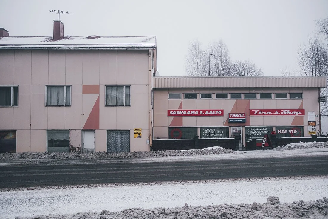 A two-story commercial building in a snowy setting, with various signs including 'Sorvaamo E. Aalto', 'Teboil', and 'Tira & Shop'.